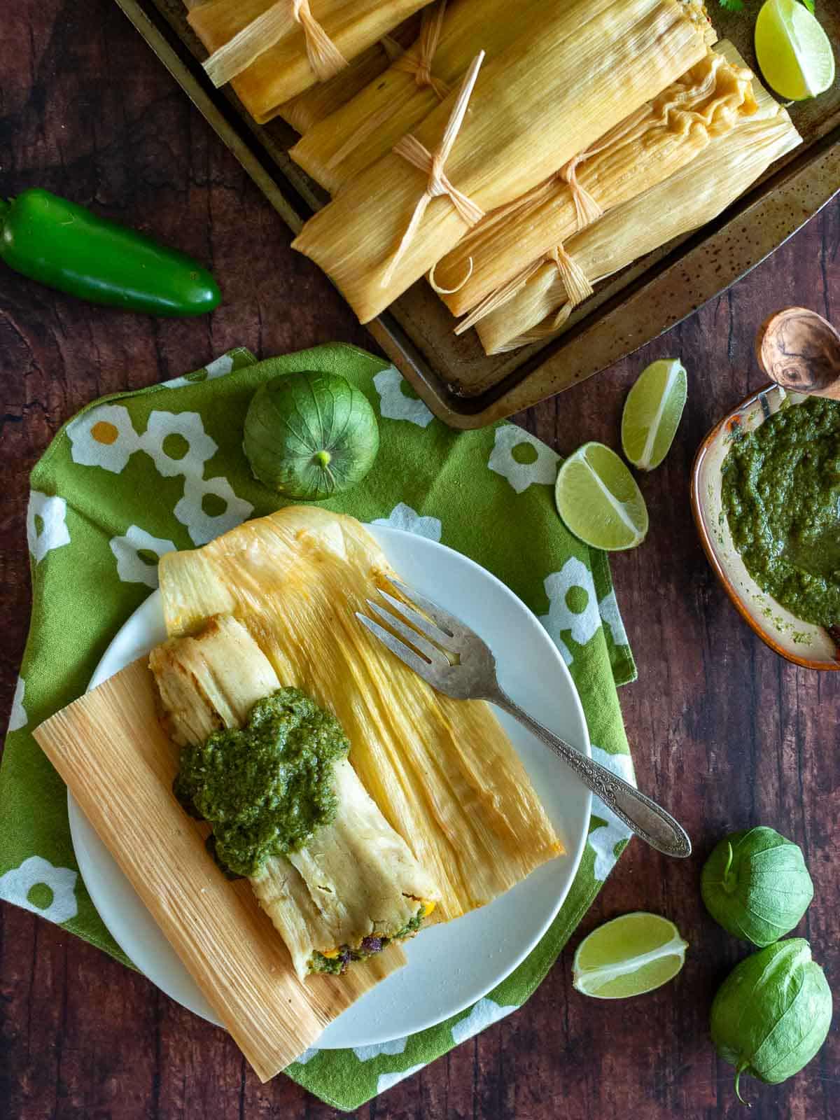 A bowl of stacked tamales on the side and in the center a freshly steamed tamal topped with tomatillo jalapeno salsa.