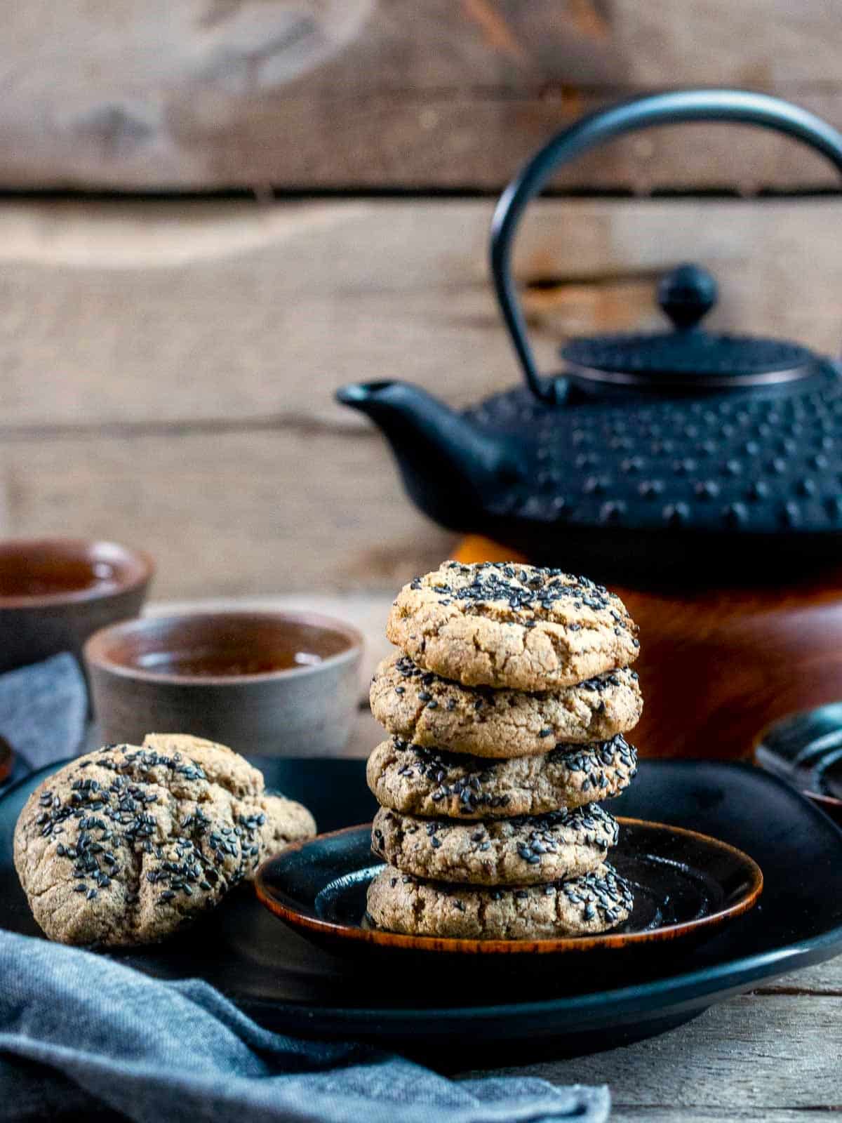 Stacked chewy black sesame cookies on a black plate with a black tea set.