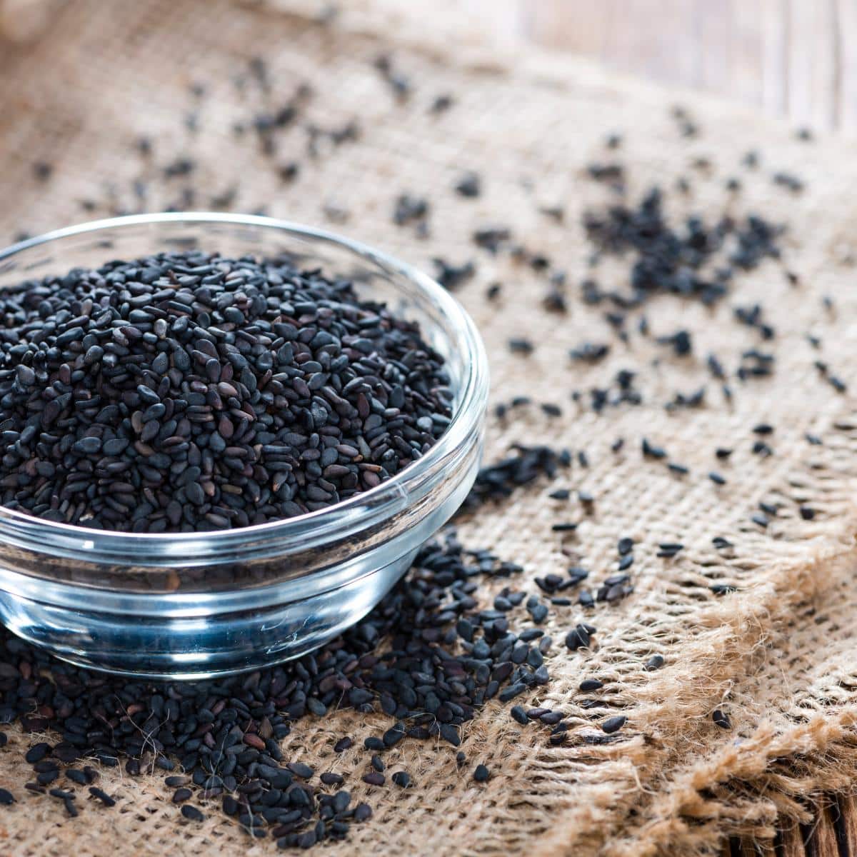 Black sesame seeds in a clear bowl scatter around on the table cloth.