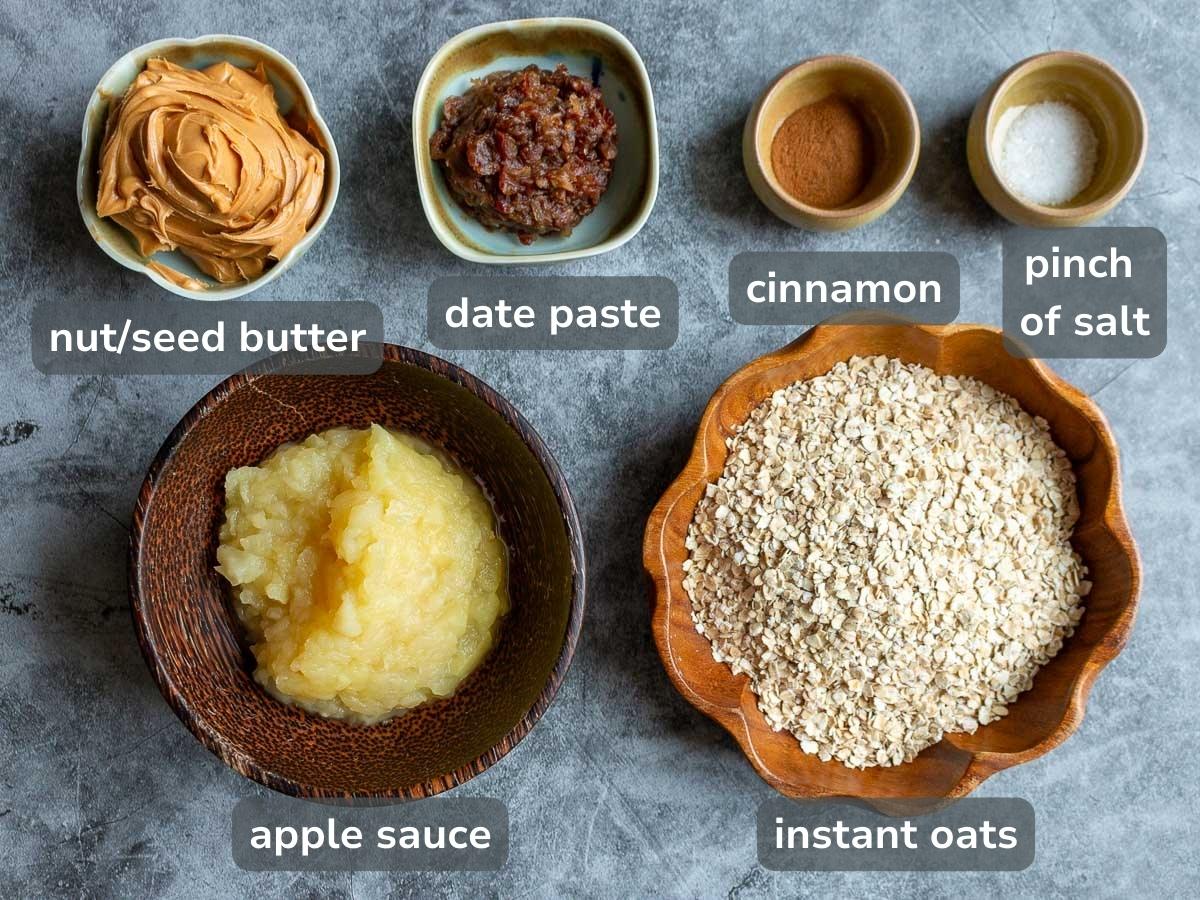 Dried oats, applesauce, date paste, nut butter, cinnamon laid out on a gray table. 