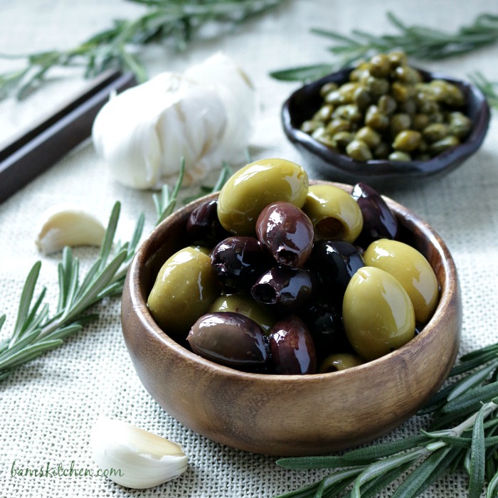 Bowl of olives, capers, garlic and rosemary on a beige cloth.