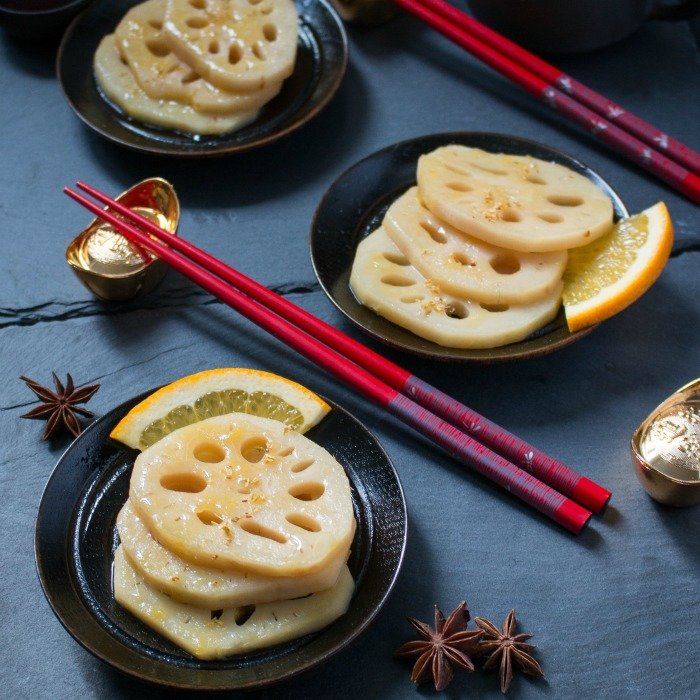 Braised orange lotus roots on black plates garnished with oranges, anise and red chopsticks.