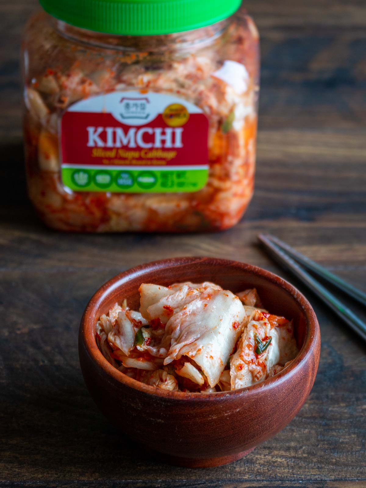 Kimchi in a wooden bowl with a container of kimchi in the background.