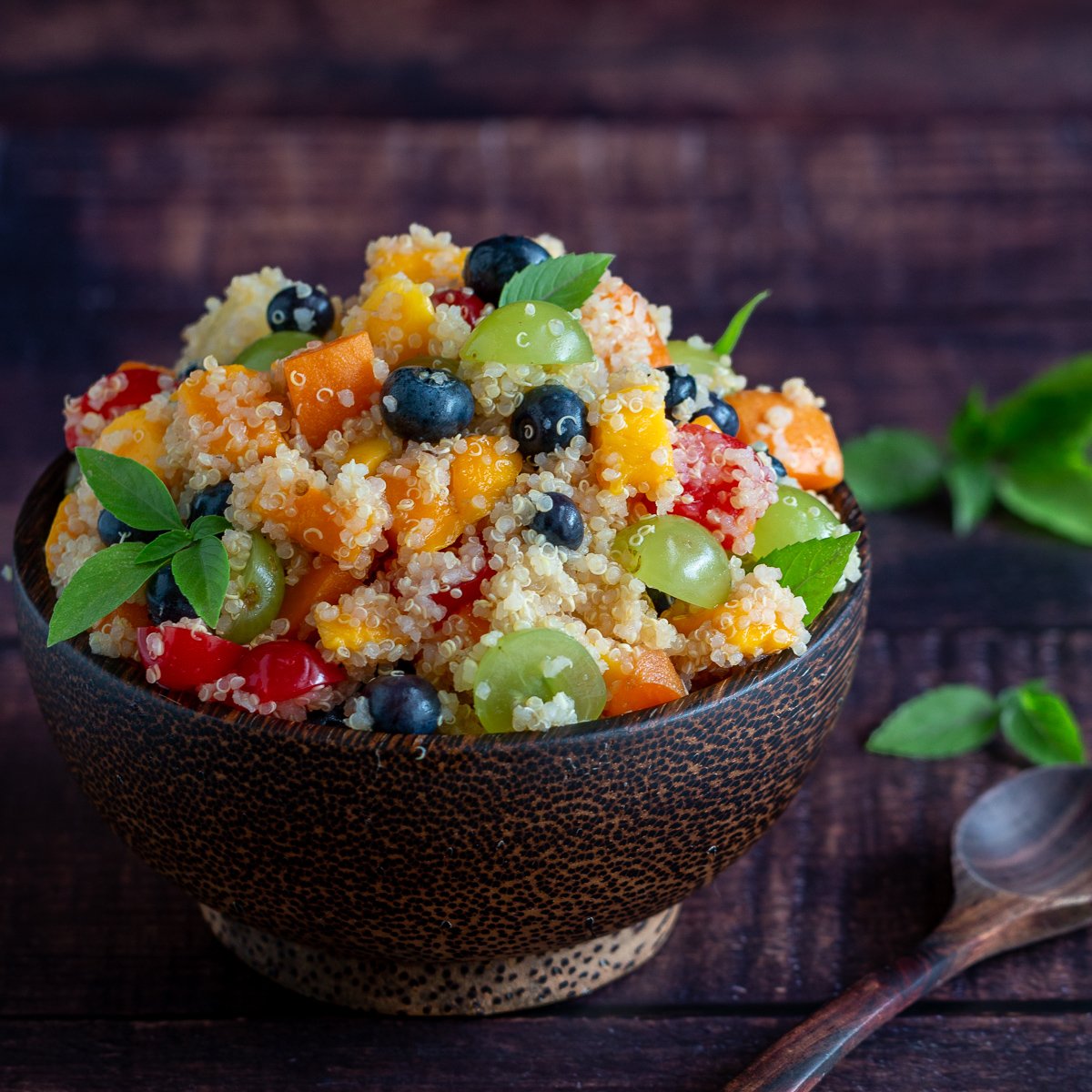 Quinoa fruit salad in a wooden bowl garnished in mint with a wooden spoon.