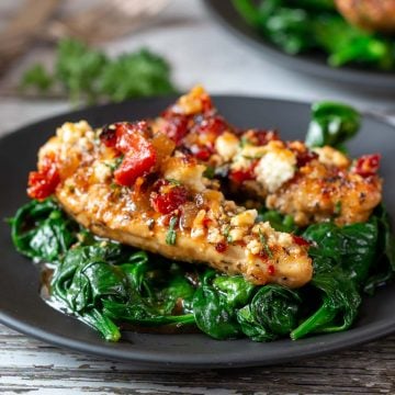 2 delicious Feta Baked chicken tenderloins over a bed of sautéed garlicky spinach on a black plate.