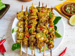 Thai pork Skewers perfectly grilled on a white plate next to a bowl of Thai basil dipping sauce.