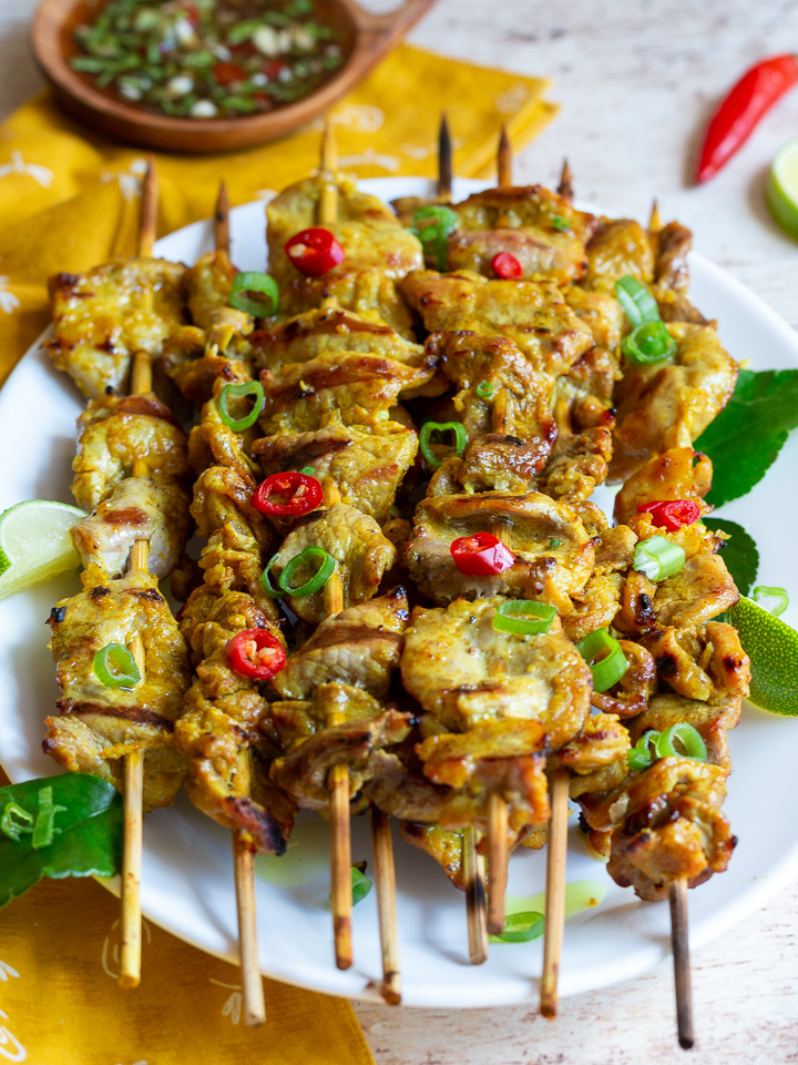 Turmeric Pork Skewers just off the grill garnished with chili and holy basil and sauce in a little white bowl. 