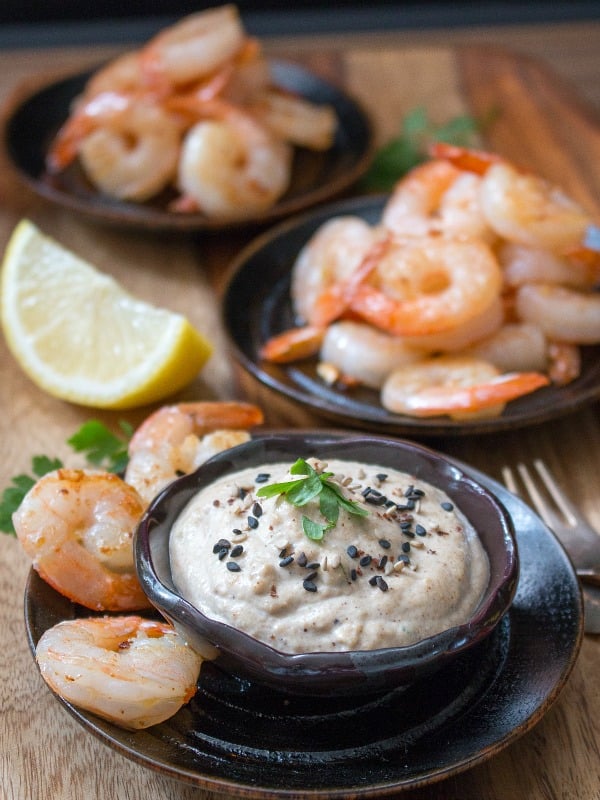 Tahini dip sprinkled with sesame seeds in a little brown pottery dish with stir fried shrimp around and a lemon slice.
