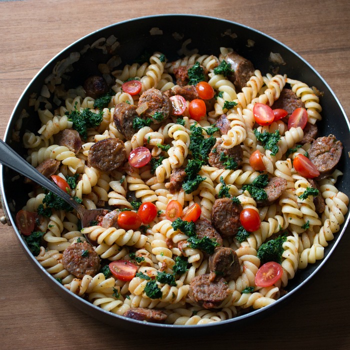 Sausage pasta tossed in a black pan with tomatoes and spinach with a spoon.