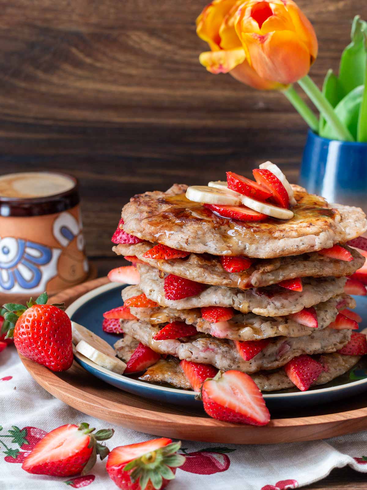 Chia seed pancakes stacked with strawberries in between and a cup of coffee and fresh tulips in the background.