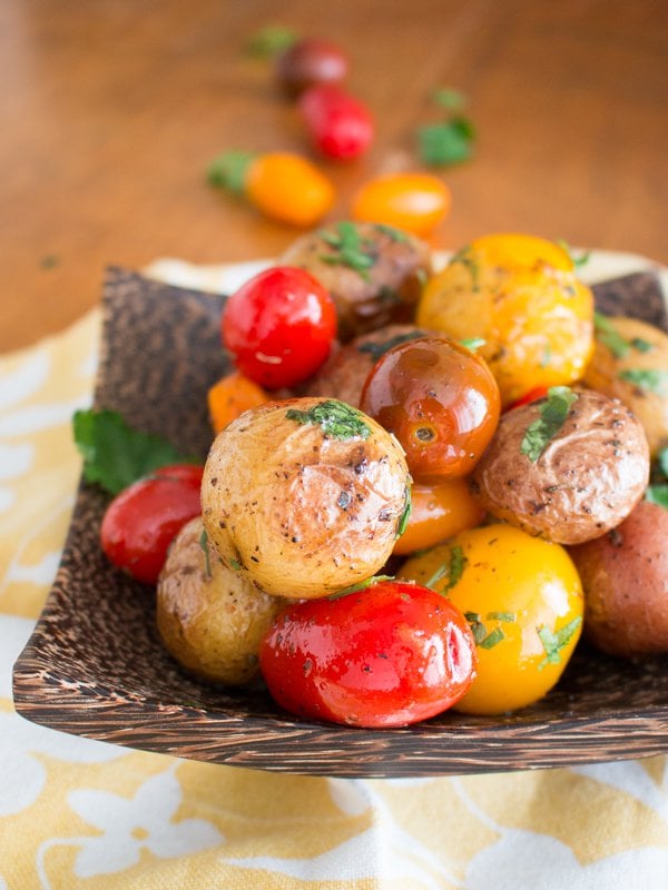 Plated on a brown wooden plate is the potatoes and tomatoes with fresh herbs sprinkled on top.