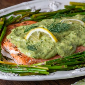 Baked salmon with avocado sauce on top with asparagus on a white serving platter.