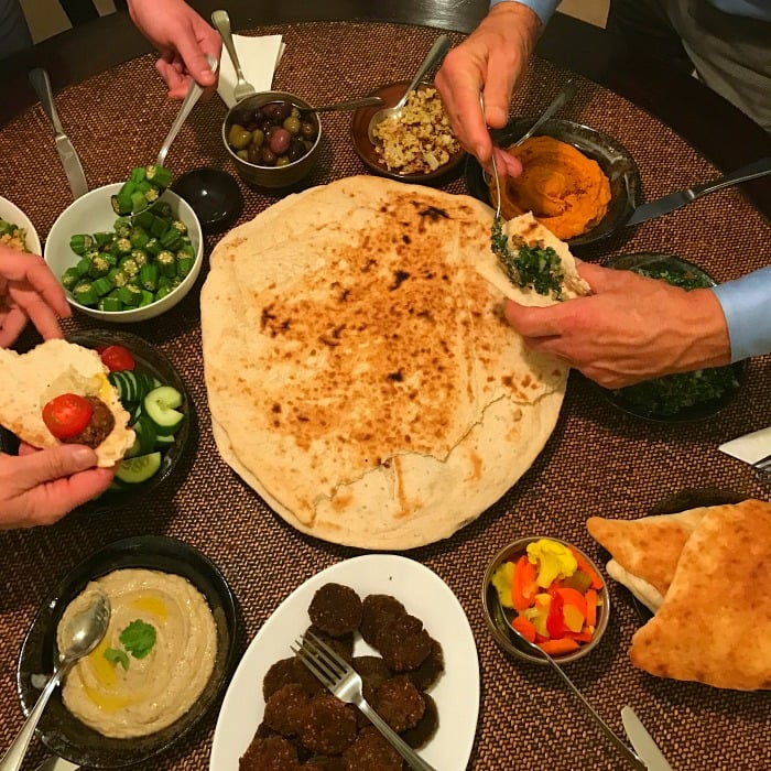 A party with 4 people grabbing flat breads, falafel, olives, tabbouli and all the toppings. HUGE flatbread in the middle and 11 delicious Middle Eastern dishes around the table.