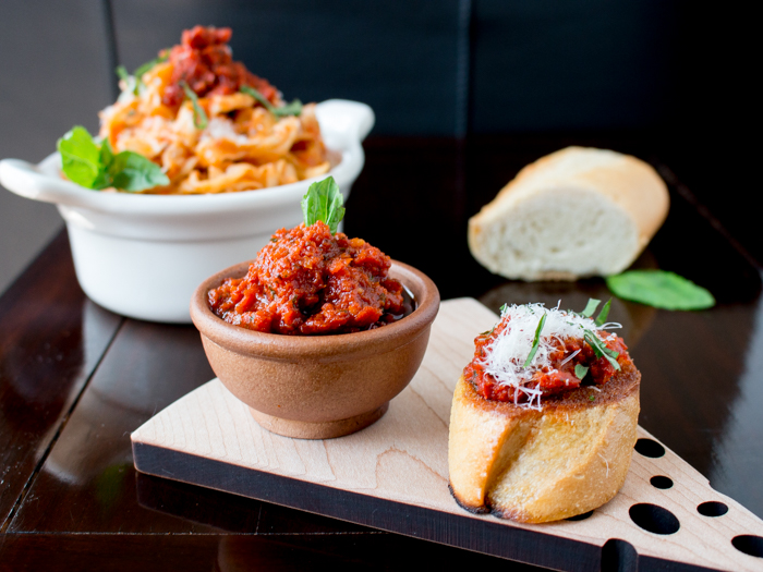 Sun-dried tomato pesto slathered over a crostini with extra pesto on the side on a wooden board shaped like a slice of cheese.