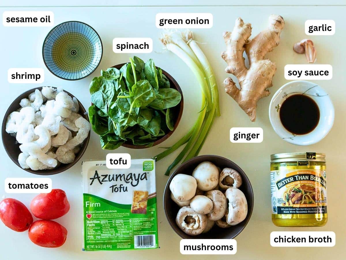 Ingredients to make Asian shrimp and tofu soup laid out on a beige board.