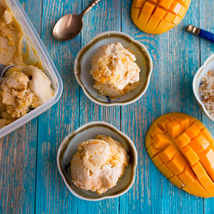 No-Churn Dairy Free Mango Swirl Ice in small cups with spoons and perfectly ripe mangoes on a blue background.
