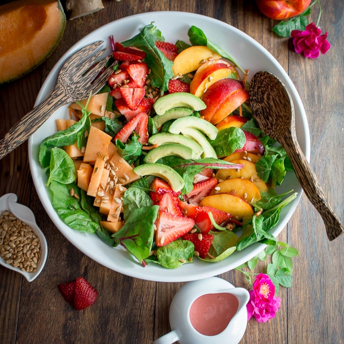 Vegan fruit salad with raspberry rose dressing in a white bowl with wooden serving spoons.