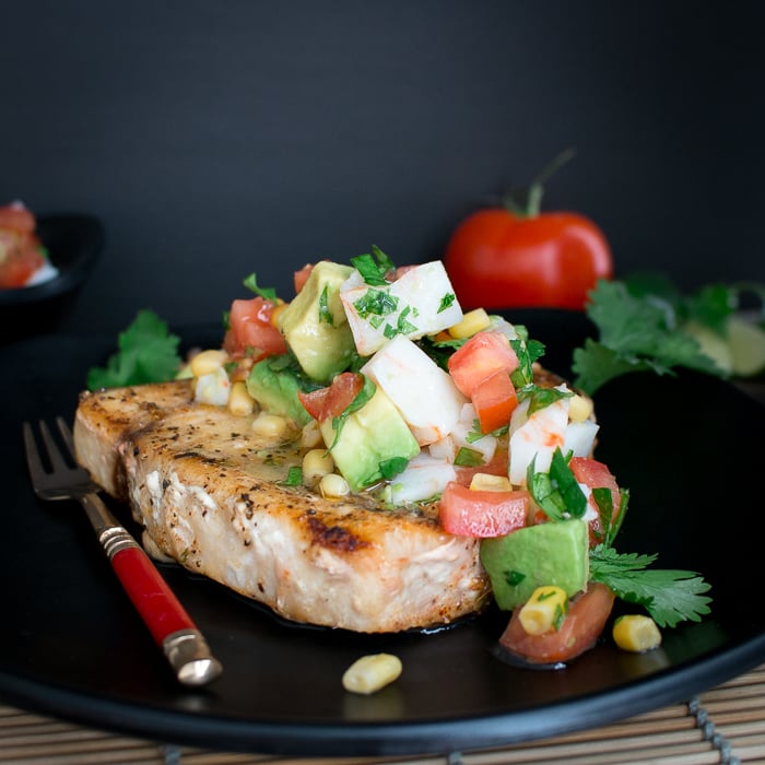 Grilled swordfish piled high with crab salsa on a black plate with a red antique fork. 