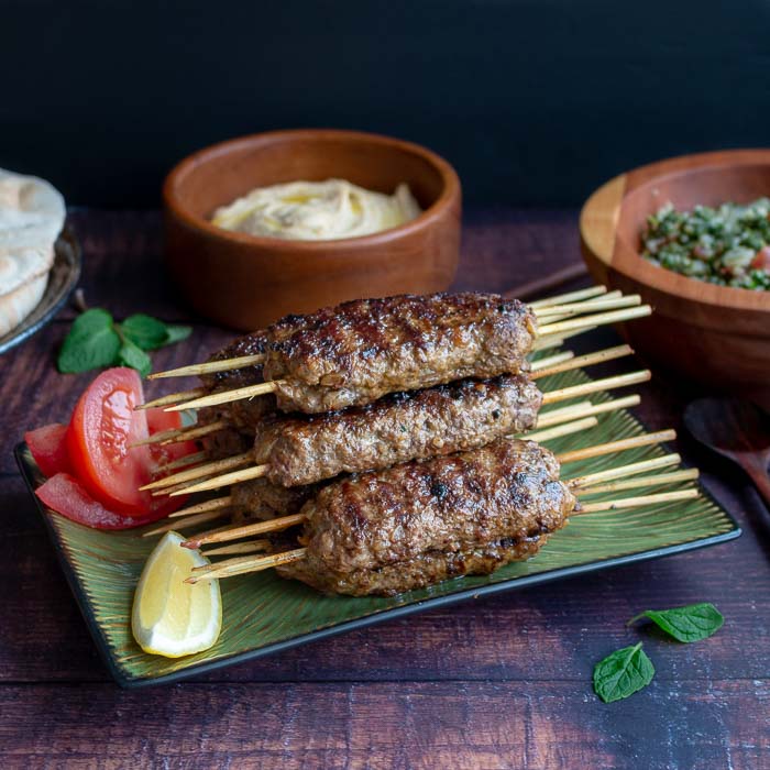 Delicious tray of grilled lamb kabobs garnished withe slices of lemons, tomatoes and pita breads and hummus in the background.