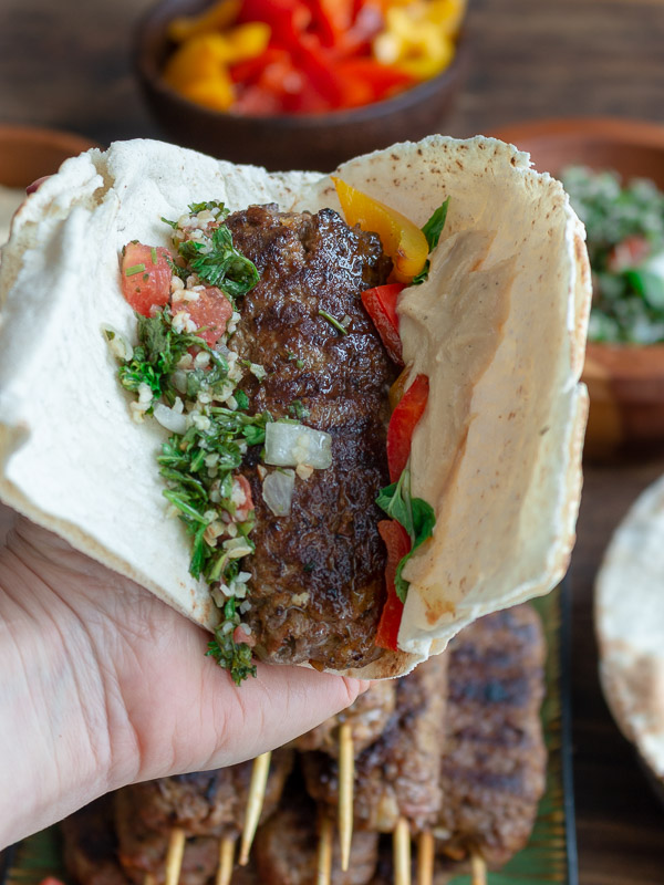 A lamb kabob held in a hand nestled in a pita bread with hummus, grilled peppers, tabbouleh and all the fixings. 