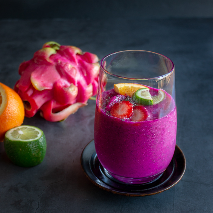 Glass of dragon fruit smoothie with a whole dragon fruit, orange, lime in the background.