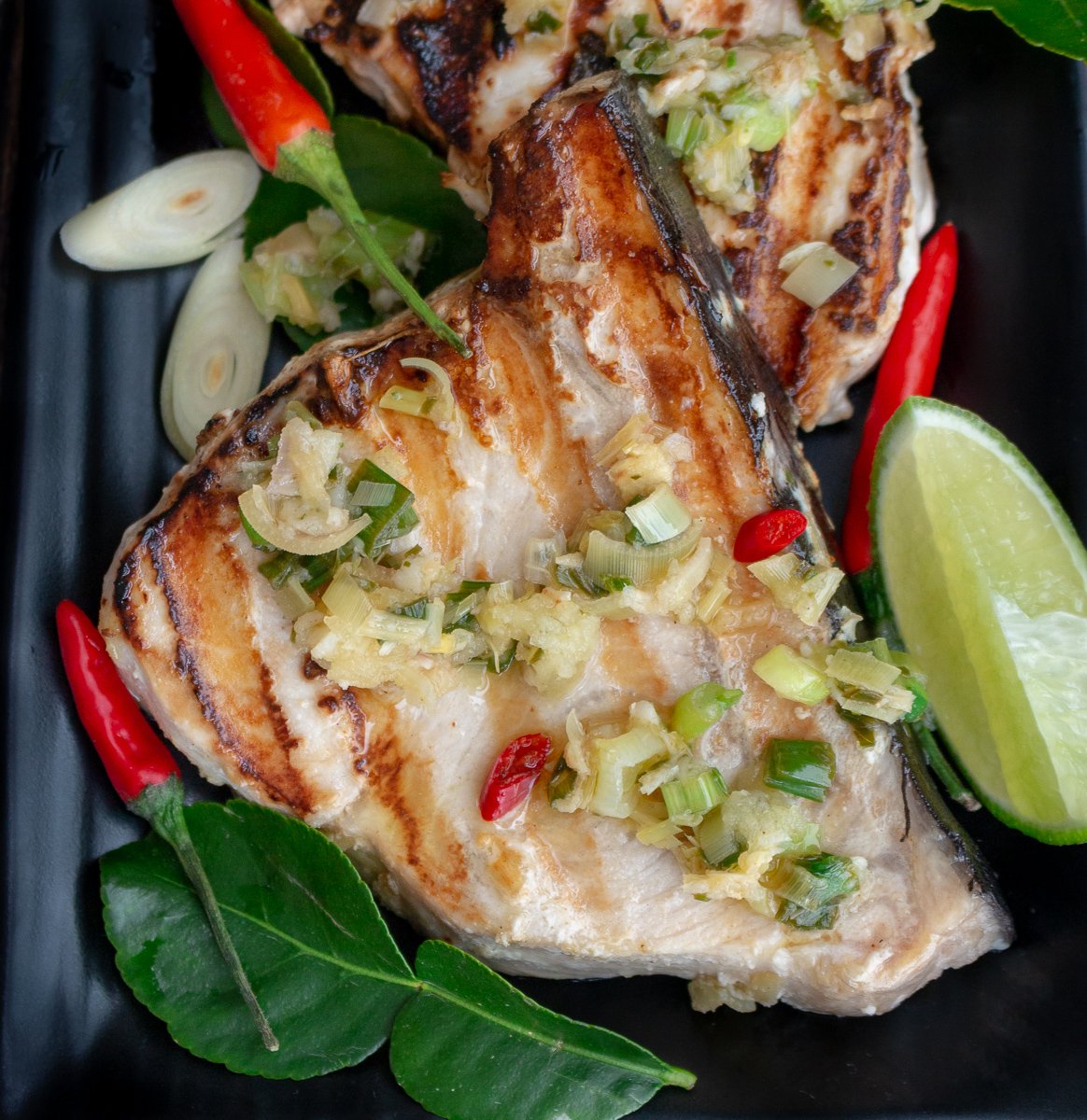Tropical Thai Swordfish just hot off the grill, topped with lemongrass sauce and chilis, and limes on a black plate with perfect grill marks.