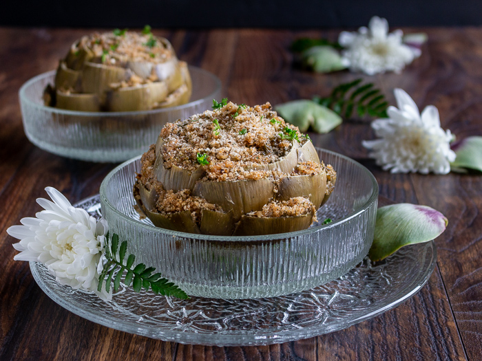 2 gigantic globe artichokes stuffed and steamed in glass bowls with white flowers around the bowls.