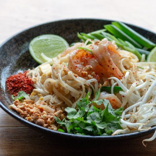 Bowl of delicious Pad Thai with all the fixings such as ground peanuts, chilis, green onions and limes on the side in a black bowl.