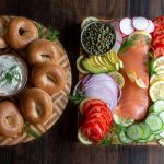 Round cutting board with bagels and cream cheese on the left and on the right a rectangle cutting board packed with lox, tomatoes, cucumbers, capers, lemons dills, radishes and onions.