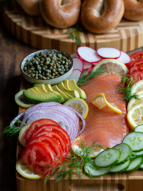 Round cutting board with bagels and cream cheese on the left and on the right a rectangle cutting board packed with lox, tomatoes, cucumbers, capers, lemons dills, radishes and onions.