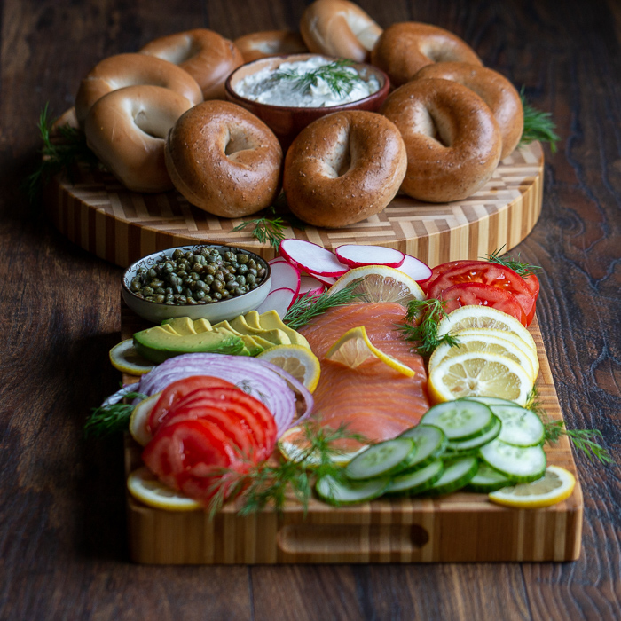 Round cutting board with bagels and cream cheese on the left and on the right a rectangle cutting board packed with lox, tomatoes, cucumbers, capers, lemons dills, radishes and onions.