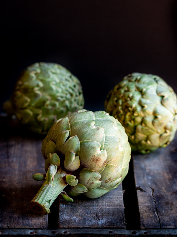 3 extra large globe artichokes on a dark wooden box with the stem facing towards you. They are whole and just home from the farmers market. 