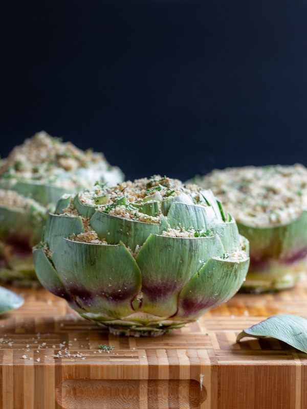 3 artichokes stuffed on a bamboo cutting board ready to be steamed.