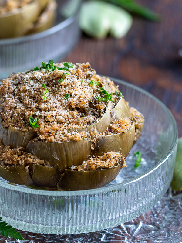 Close up of gigantic whole stuffed and steamed globe artichokes sprinkled with a little extra parmesan and fresh parsley in a glass bowl surrounded by white flowers. 