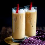 Two glasses of Easy Coconut Iced Chai Tea Latte with red straws and lots of ice on a wooden board with a polka dot napkin.