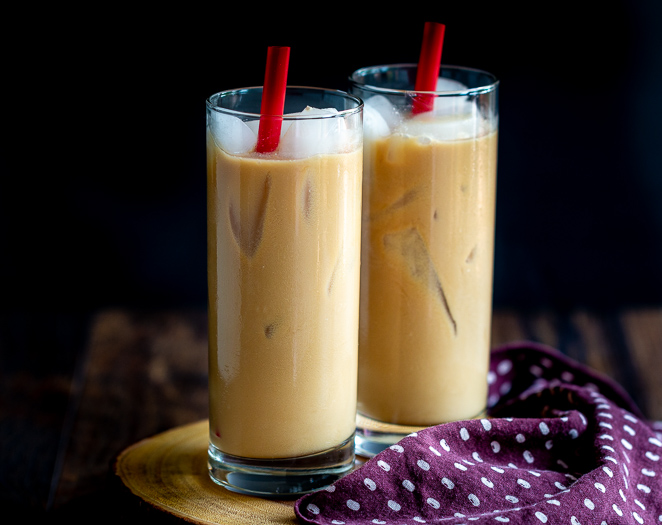 Two glasses of Easy Coconut Iced Chai Tea Latte with red straws and lots of ice on a wooden board with a polka dot napkin.