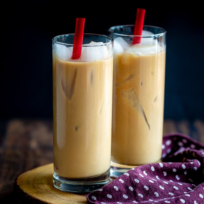 Two glasses of Easy Coconut Iced Chai Tea Latte with red straws and lots of ice on a wooden board with a polka dot napkin.