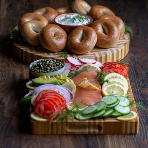 DIY Ultimate Bagel Bar Brunch on a cutting board with lox, cream cheese, vegetables and all the fixings and a platter of bagels.
