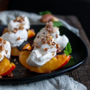 Grilled Nectarines with Coconut Cream on a black plate garnished with crushed nuts and mint leaves.