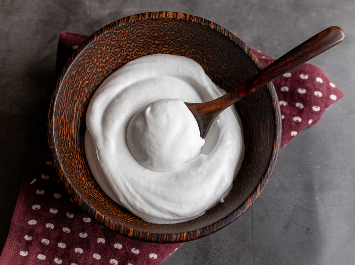 Top down photo of creamy dreamy coconut whipped topping in a brown wooden bowl with a dollop in a wooden spoon.