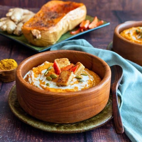 Roasted butternut squash and carrot soup with ginger in a wooden bowl topped with crispy croutons and pepitas.