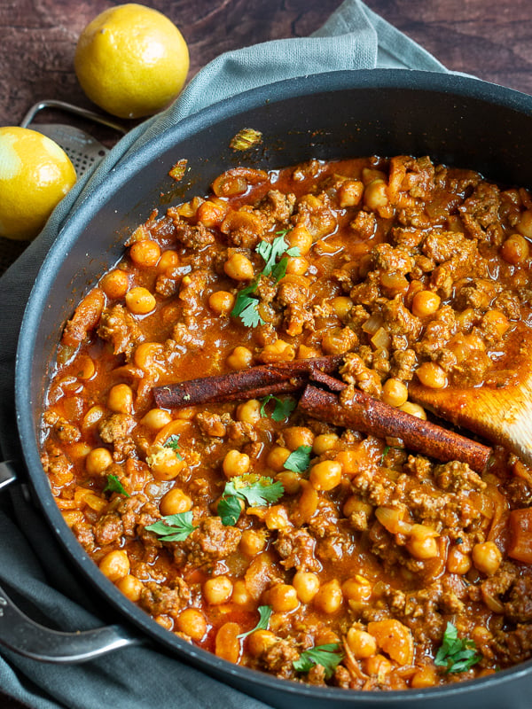 Moroccan stew in a black pan being stirred with a wooden spoon and the whole cinnamon sticks still inside.