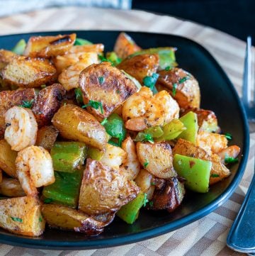 Piled in a black bowl is roasted crispy potatoes, green peppers, onions and shrimp.