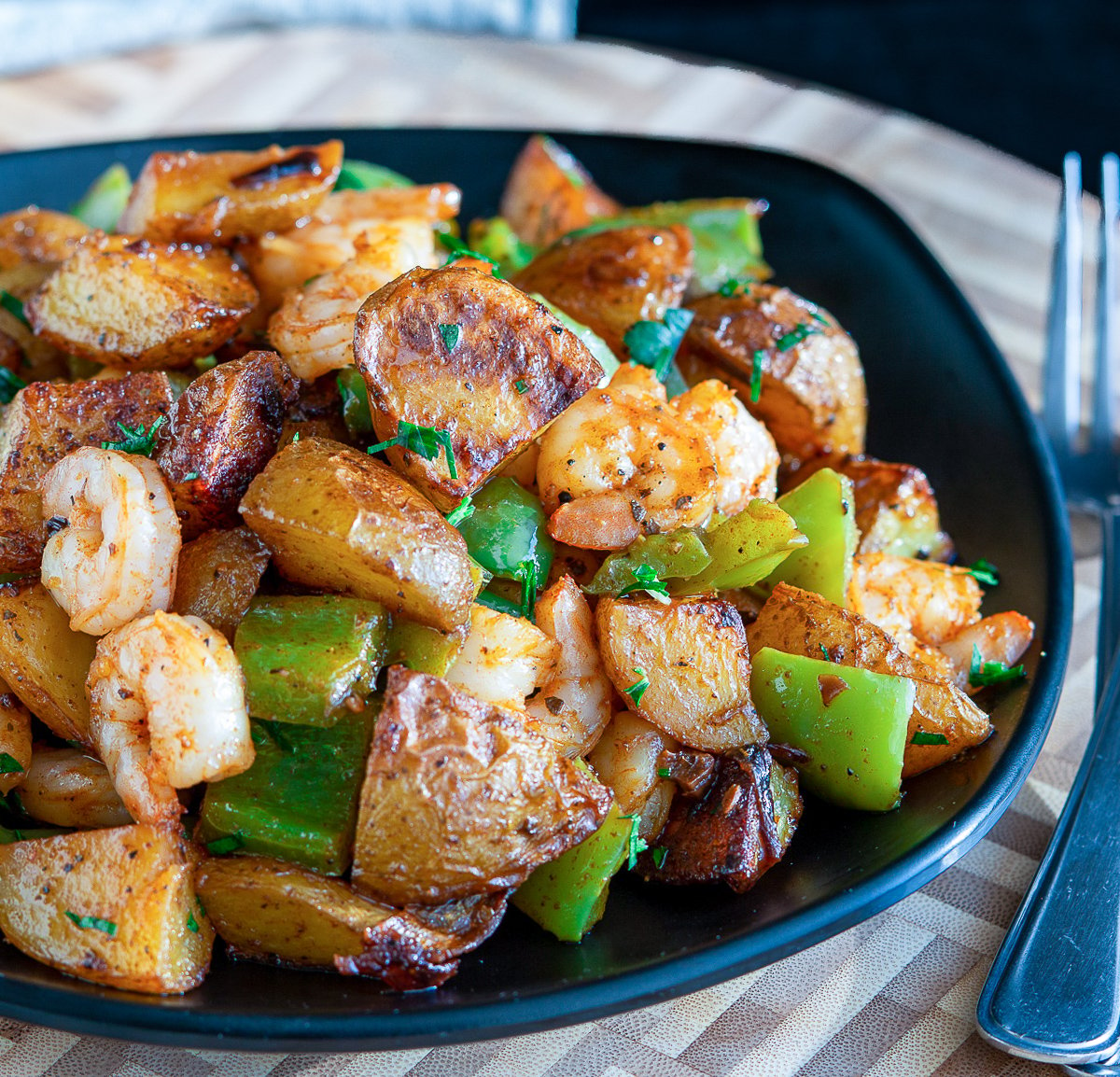 Piled in a black bowl is roasted crispy potatoes, green peppers, onions and shrimp.