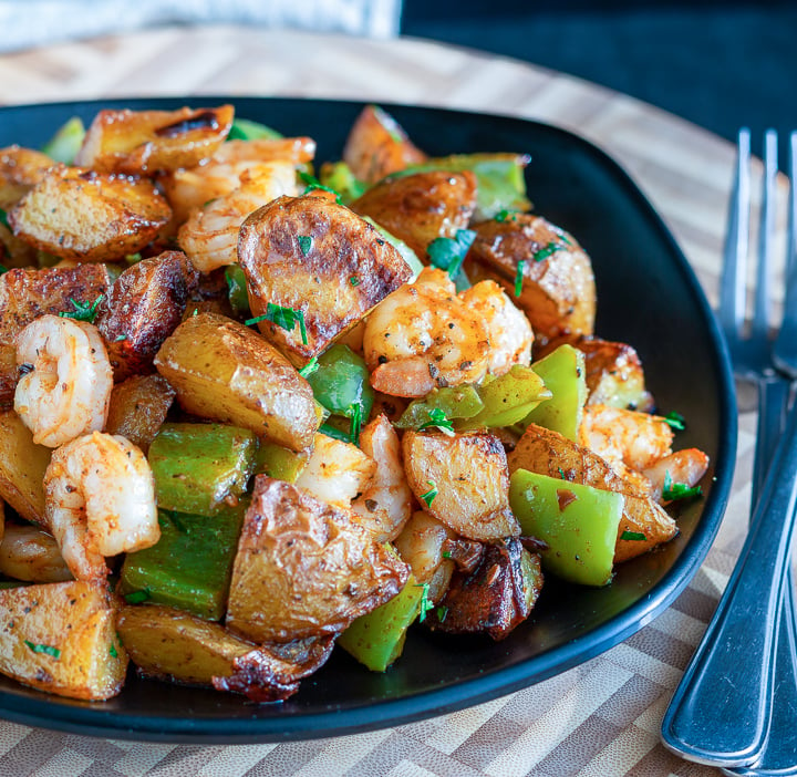 Piled in a black bowl is roasted crispy potatoes, green peppers, onions and shrimp.