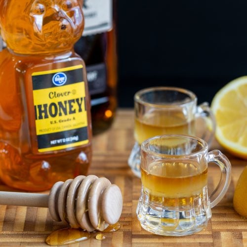 2 shots of Whiskey Honey Lemon Cough Syrup with a bottle of honey and whiskey and a half a lemon on a cutting board.