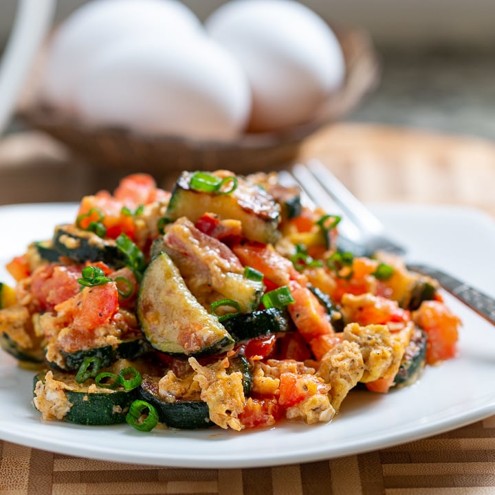 Delicious bowl of Italian Scrambled Eggs on a white plate with a fork ready to eat and a cup of coffee and whole eggs in the background.