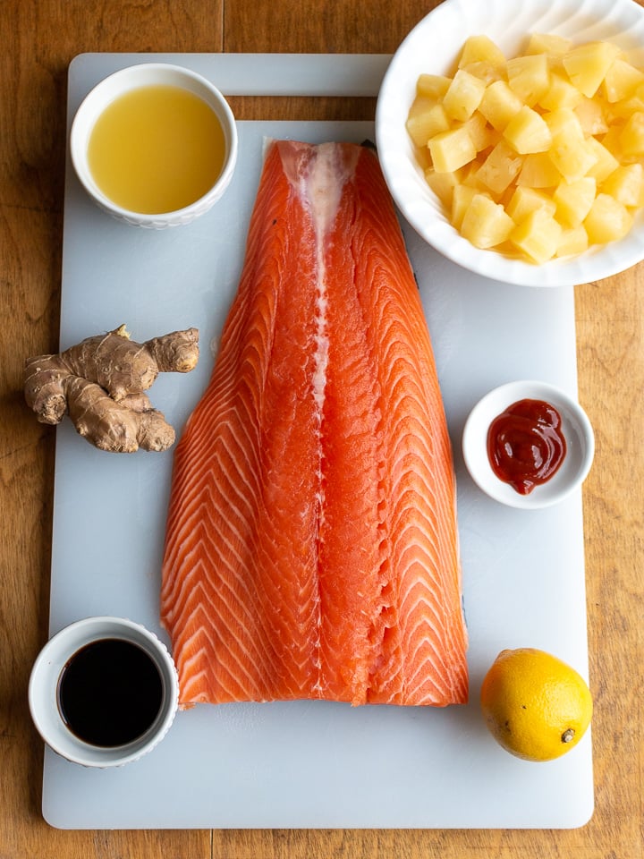 Top down phone of salmon fillet with pineapple, sriracha sauce, lemon, teriyaki sauce, fresh ginger and pineapple juice around the cutting board.