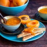 Toast slathered in apricot freezer jam with a bowl of jam next to it and fresh apricots in the background on a turquoise plate.