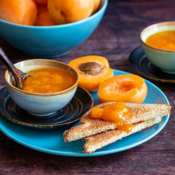 Toast slathered in apricot freezer jam with a bowl of jam next to it and fresh apricots in the background on a turquoise plate.